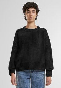 Zwarte gebreide trui met een geribbelde textuur, brede halslijn en verlaagde schouders. Gecombineerd met lichtblauwe versleten jeans.
