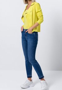 Żółta, teksturowana marynarka na żółtej bluzce z koronkowym wykończeniem, w połączeniu z dopasowanymi niebieskimi dżinsami i białymi sneakersami, na neutralnym tle.