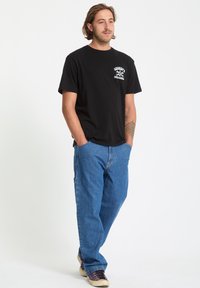 Czarny bawełniany t-shirt z białą grafiką na piersi, w połączeniu z niebieskimi spodniami cargo z denimu i fioletowymi trampkami na kremowej podeszwie.