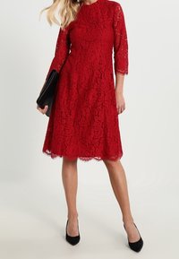 Punainen pitsimekko, jossa on kolme neljännestä hihoista, pyöreä kaula-aukko, aallotettuhelma ja tiukka siluetti. Yhdistetty mustiin korkokenkiin ja clutch-laukkuun.