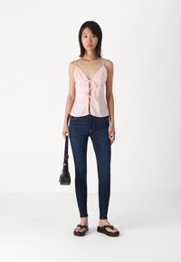 Camiseta de satén rosa con escote en V profundo y detalle de lazo en el frente, combinada con jeans skinny de color azul oscuro y sandalias de playa negras. Pequeño bolso negro.