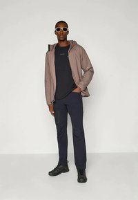 Muškarac u bež dukserici s kapuljačom preko crne majice i hlača, s crnim cipelama i naočalama s bijelim okvirom, stoji naslonjen na svijetlosivu pozadinu.