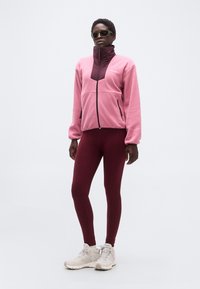 Γυναίκα με γυαλιά ηλίου, ροζ-βαθύ ροζ μπουφάν με φερμουάρ, βαθύ ροζ κολάν και μπεζ παπούτσια πεζοπορίας, στέκεται μπροστά σε απλό φόντο.