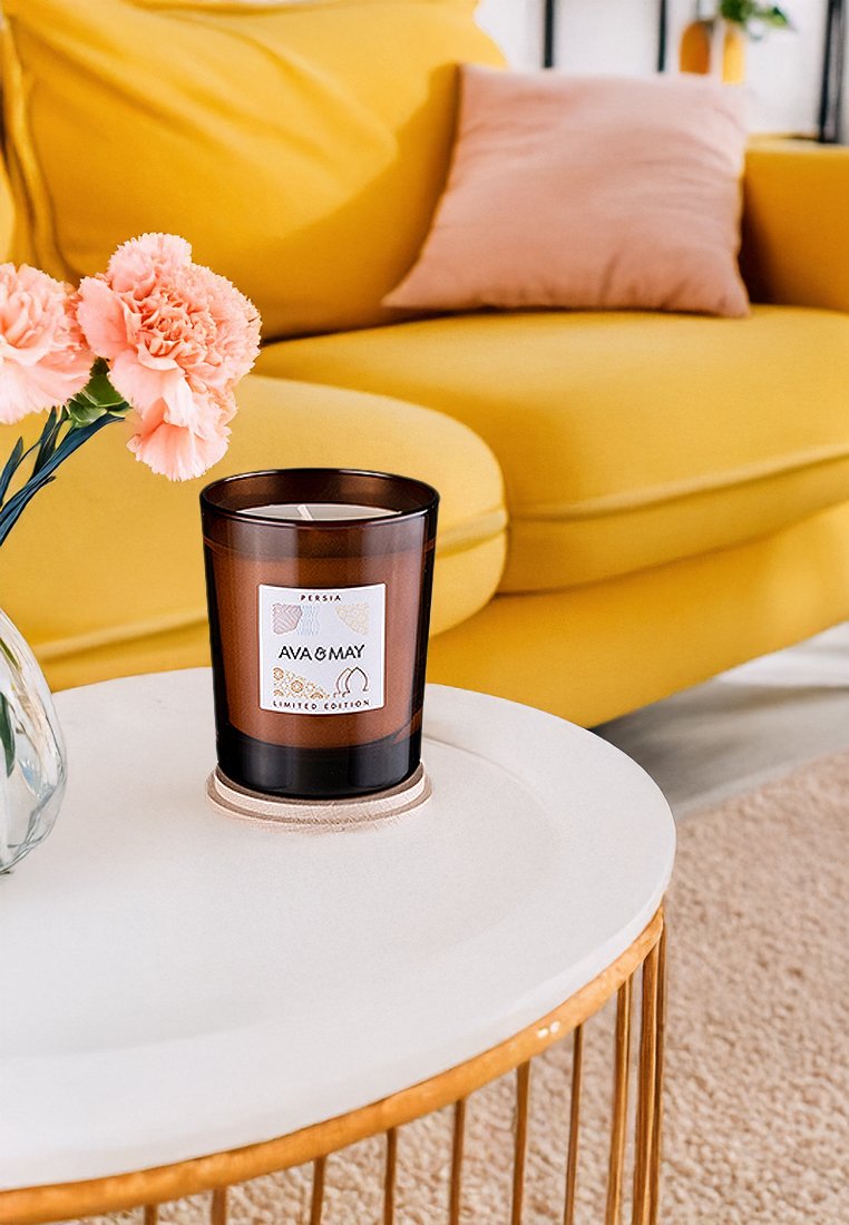 Bernsteinfarbene Glaskerze mit einem weißen Etikett, auf dem "Ava & May" steht. Auf einem runden weißen Tisch platziert, neben einer Vase mit rosa Blumen und einem gelben Sofa.