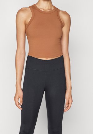 Frau trägt ein ärmelloses braunes Croptop und hoch taillierte schwarze Leggings vor einem einfarbigen hellen Hintergrund.