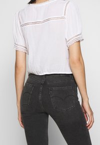 Weißes kurzärmeliges Oberteil mit Spitzenbesatz, kombiniert mit dunkelgrauen engen Jeans, die über zwei Gesäßtaschen und eine klassische Denimtextur verfügen.