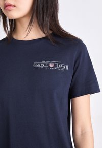T-shirt en coton bleu marine à manches courtes, avec un texte imprimé "GANT 1949" et un logo. Design simple à col rond. Texture douce.