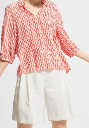 Kvinde iført løse hvide shorts og en rød mønstret bluse med trekvart-ærmer mod en ensfarvet baggrund.