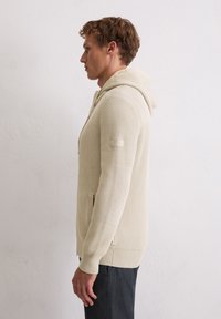 Beige gestrickter Hoodie mit strukturiertem Finish, ausgestattet mit einer Kapuze, Fronttaschen und gerippten Bündchen. Seitenschlitze am Saum für mehr Bewegungsfreiheit.