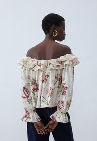 Blouse à épaules dénudées en tissu crème avec motif floral, dotée de poignets et d'un col à volants, associée à un denim bleu foncé.