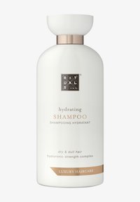 Bouteille de shampoing blanche avec une forme arrondie, des accents dorés et une texture mate. L'étiquette présente du texte dans une police élégante, indiquant une hydratation pour cheveux secs.