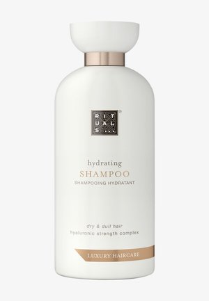 Weiße Shampoo-Flasche mit runder Form, goldenen Akzenten und mattem Finish. Das Etikett zeigt einen Text in eleganter Schrift, der auf eine Hydratation für trockenes Haar hinweist.