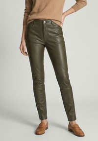 Vrouw draagt donkergroene leren broek met een beige trui en tankleurige loafers, staand met één hand op haar heup tegen een effen achtergrond.