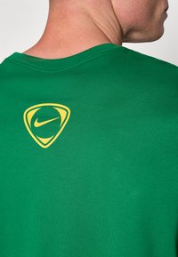 Grøn bomulds t-shirt med et gult Nike-logo i skjoldform på ryggen. Rund halsudskæring og korte ærmer med en glat tekstur.