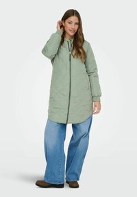 Quiltēts zaļš garš mētelis ar rāvējslēdzēju, sānu kabatām un kapuci, uzvilkts pār brīvi pieguļošām ziliem džinsiem un brūniem potīšu zābakiem.