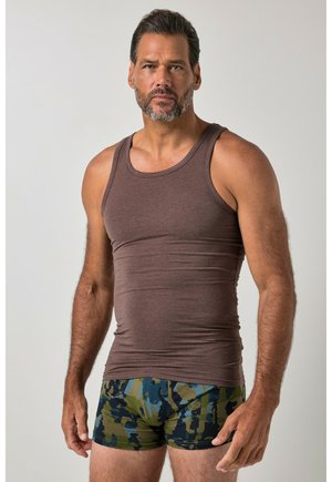 Hombre con barba que lleva una camiseta marrón sin mangas y pantalones cortos de camuflaje azul y verde, de pie frente a un fondo sencillo.