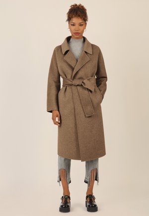 Brown woolen coat with a belted waist, wide lapels, and long sleeves, paired with distressed light blue jeans and black platform shoes.