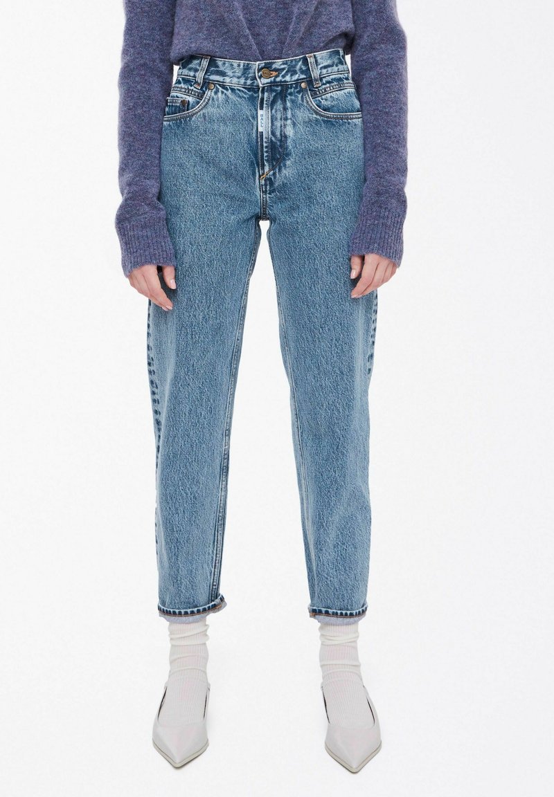 Høy midje blå denimjeans med avsmalnende ben, subtile slitasjer og sytt detaljer ved mansjettene, kombinert med hvite ribbede sokker og grå spisse sko.