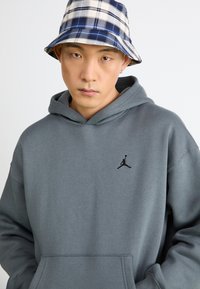 Grå hættetrøje med et sort Jumpman-logo på brystet, med hætte og frontlomme. Båret med en blå og hvid ternet bøttehat.
