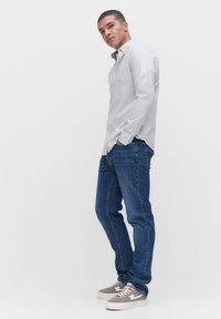 Lichtgrijs knoopshirt, blauwe spijkerbroek en grijze sneakers. Shirt heeft lange mouwen en een borstzak. Casual outfit, staande pose.