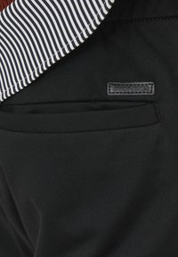 Pantaloni neri con una texture liscia, dotati di una tasca posteriore e di un'etichetta loggata rettangolare nera. Patta a righe visibile in alto.