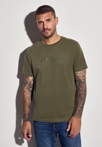 Muž s krátkými vlasy a tetovanými pažemi, který má na sobě olivově zelené tričko "Crafted Goods" a modré džíny, na jednobarevném světlém pozadí.