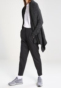 Tmavě šedý pletený šál přes bílou košili se zapínáním na knoflíky, v kombinaci s vysokými černými kalhotami a světle šedými teniskami s bílými detaily.