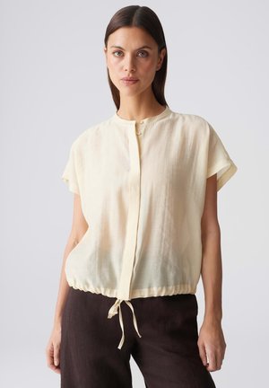Femme aux cheveux bruns et lisses porte un chemisier ample crème à manches courtes avec un cordon à la taille et un pantalon foncé sur fond uni.