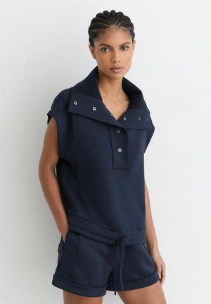 Kvinde med flettet hår iført navy ærmeløs top med trykknapper og matchende shorts med snøre, stående foran en hvid væg.