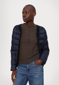 Navy puffer jacket with a lightweight design and quilted texture, worn over a brown henley sweater and blue jeans.