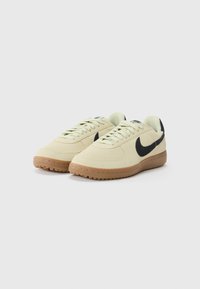 Beige sneakers med en blød ruskindsoverdel, sort swoosh-logo og gummisål; har runde snørebånd og et minimalistisk design.
