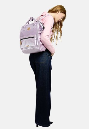 Junge Frau in einem pinkfarbenen Pullover und dunklen Jeans, die einen glänzenden lavendelfarbenen Rucksack trägt, steht im Profil mit den Händen in den Taschen.