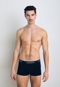 Navy blue boxer briefs with a soft texture and white "LACOSTE" logo on the waistband. Model has an athletic build and standing posture.