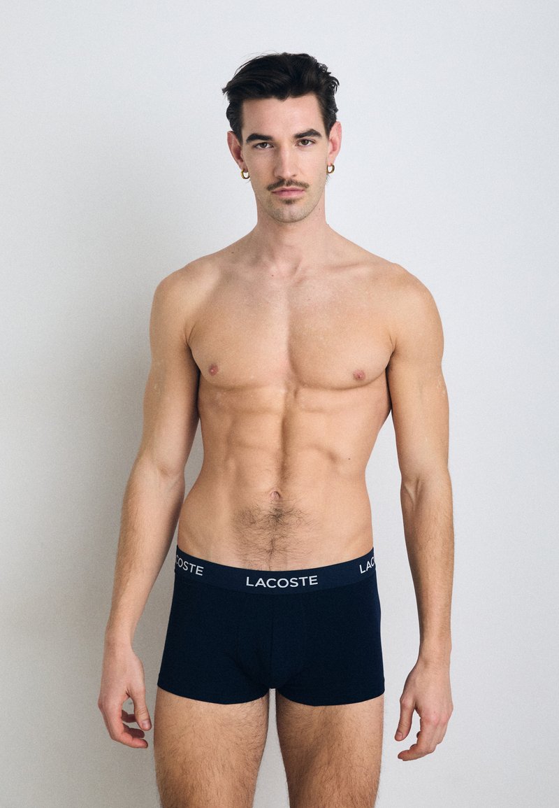 Granatowe bokserki z miękkiego materiału z białym logo "LACOSTE" na pasku. Model ma atletyczną budowę ciała i stoi w wyprostowanej pozycji.