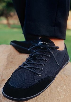 Zapatilla negra de cordones con la marca "GROUNDIES" usada en un pie apoyado sobre una roca, con pantalones negros visibles.