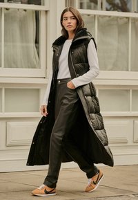 Vestă lungă neagră cu umplutură, cu glugă, având un design matlasat. Purtați-o peste o cămașă albă cu mânecă lungă și pantaloni negri din piele ecologică, asortați cu adidași bej.