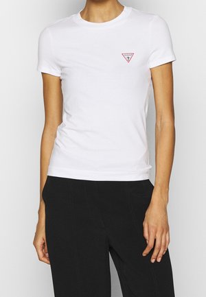 Kobieta ubrana w dopasowany biały t-shirt z krótkim rękawem z małym czerwonym logo Guess w kształcie trójkąta na lewej piersi oraz czarne spodnie.