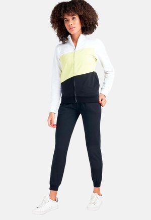 Une femme porte une veste zippée à blocs de couleurs, un pantalon de jogging noir et des baskets blanches sur un fond blanc uni.
