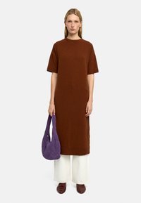 Brązowa dzianinowa sukienka oversize z krótkimi rękawami, w połączeniu z szerokimi białymi spodniami i ciemnobrązowymi butami. Trzyma fioletową plecioną torbę.