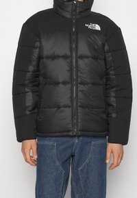 Chaqueta de plumas negra con cuello alto, cierre de cremallera y logo en el lado izquierdo del pecho. Presenta un diseño de textura acolchada y color sólido.