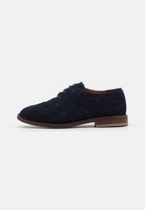 Chaussure brogue en daim bleu foncé avec design wingtip, détails perforés, semelle marron et lacets noirs, vue de côté sur fond blanc.