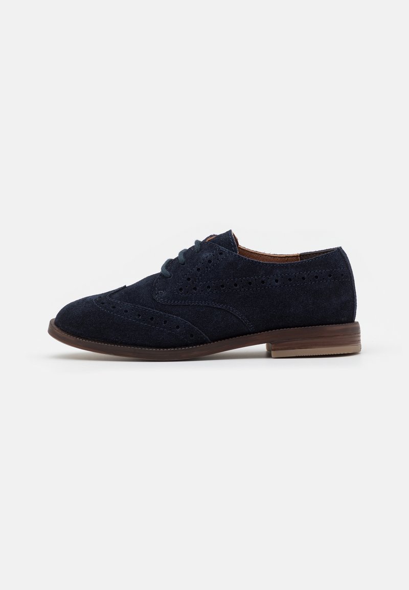 Chaussure brogue en daim bleu foncé avec design wingtip, détails perforés, semelle marron et lacets noirs, vue de côté sur fond blanc.