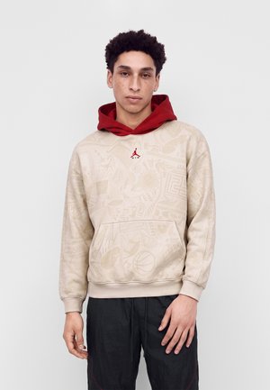 Jeune homme aux cheveux bouclés portant un sweat à capuche beige à motif avec une capuche rouge et un pantalon noir, debout devant un fond clair uni.