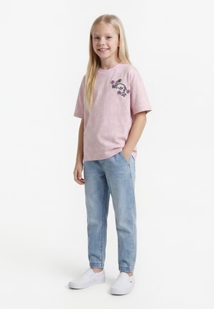 Smilende pige med langt blondt hår iført en lyserød T-shirt med blomsterprint, lyseblå jeans med elastiske manchetter og hvide slip-on sko.