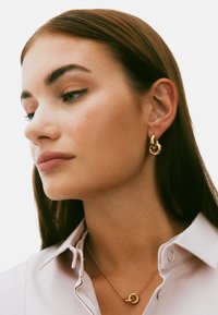 Gold hoop earrings and a gold chain necklace with a pendant, worn by a person with medium brown hair, set against a white background.