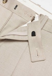 Gros plan sur la ceinture d’un pantalon beige montrant une fermeture par bouton, une patte intérieure avec crochet noir, des passants de ceinture et la texture visible du tissu.