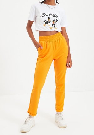 Personne portant un t-shirt blanc court avec des canards dessinés et un pantalon orange taille haute avec des baskets, debout devant un fond uni.