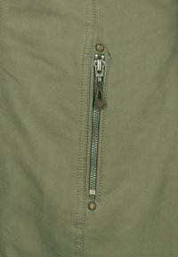 Olivenfarvet stof med en sidelukning med metalhardware og kontrasterende syninger. Glat tekstur med en subtil glans.