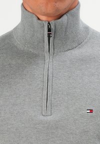 Mann trägt einen grauen, gerippten Pullover mit hohem Kragen und einem kurzen Reißverschluss, auf der Brust befindet sich ein kleines rechteckiges Logo in Rot, Weiß und Blau.
