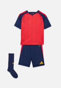 Κόκκινη αθλητική φανέλα με μπλε σκούρα μανίκια και κάθετες ρίγες, μπλε σκούρα σορτς με κίτρινο λογότυπο, και μπλε σκούρες κάλτσες με κίτρινη λεπτομέρεια, απλωμένα επίπεδα.